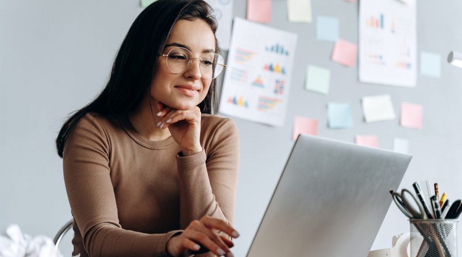 Image of young pleased, happy, beautiful business woman sit indoors in office using laptop computer. The girl wears glasses.